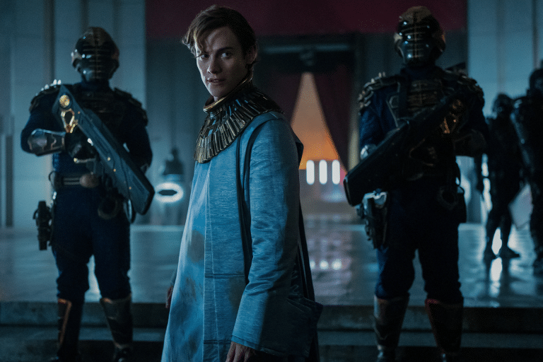 A person in futuristic clothing stands in a dimly lit hall, flanked by two armored guards with large weapons. With a blue tint and an air of intensity, the scene evokes the high-stakes drama found in Apple TV+’s diverse hits.