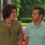 Two men stand outdoors facing each other. One wears a green shirt with layered necklaces, the other a light blue polo. Both have serious expressions, surrounded by greenery and colorful furniture—a scene fit for Apple TV+ summer streaming.
