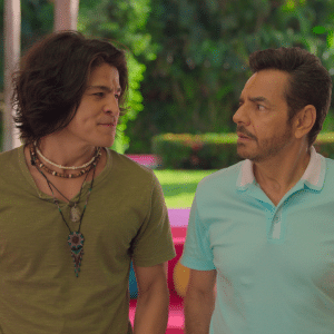 Two men stand outdoors facing each other. One wears a green shirt with layered necklaces, the other a light blue polo. Both have serious expressions, surrounded by greenery and colorful furniture—a scene fit for Apple TV+ summer streaming.