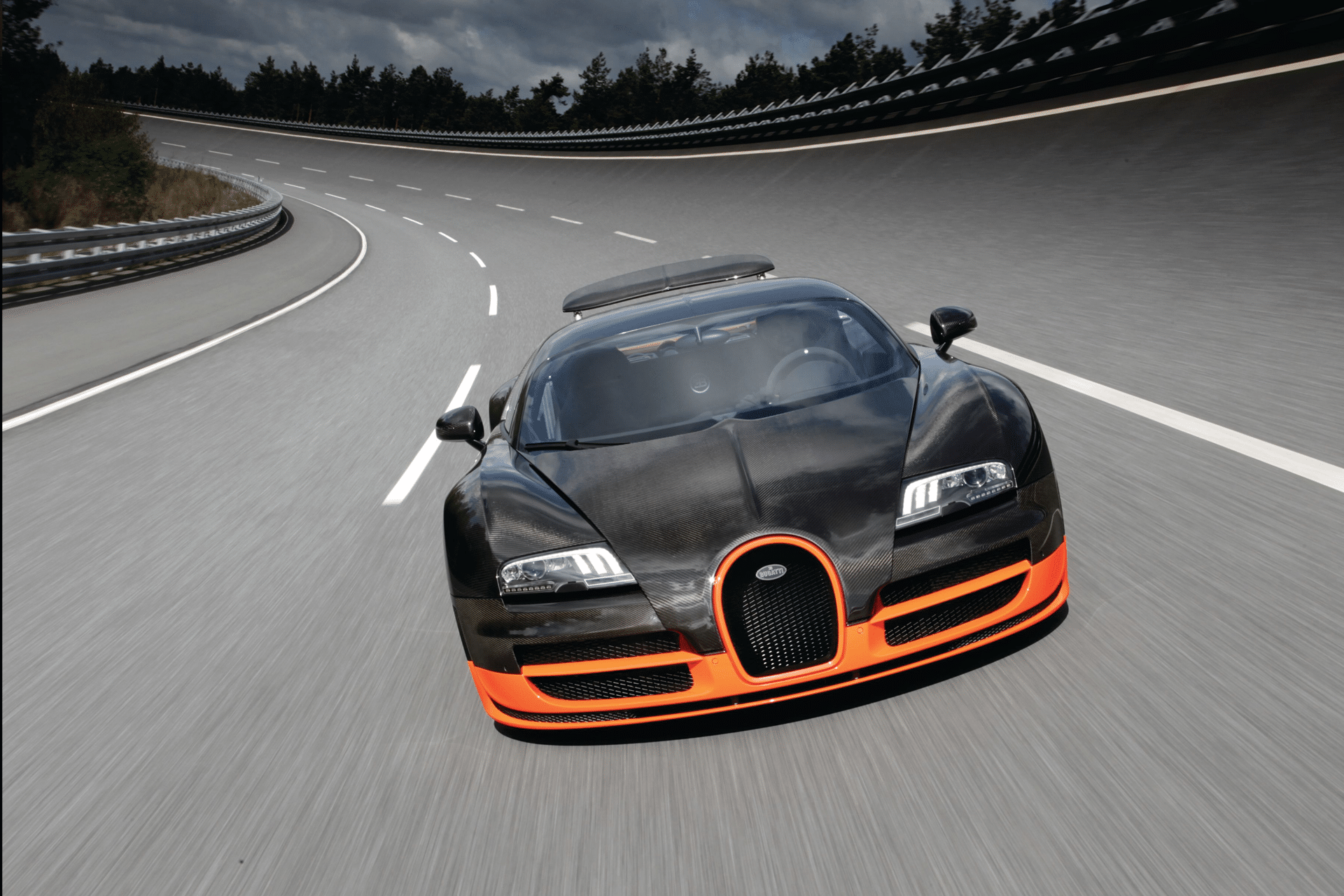 A black and orange dreame sports car speeds down an empty, curved highway with a forested area and cloudy sky in the background.