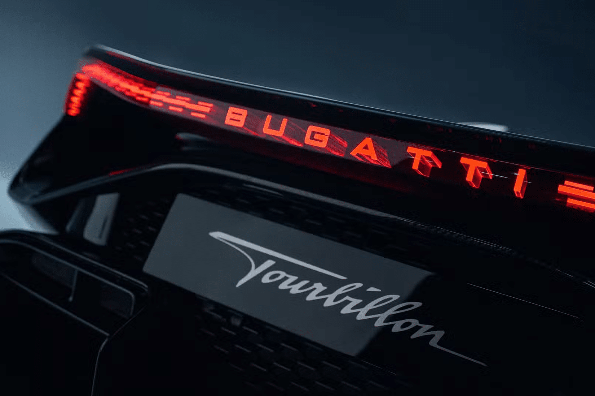 Close-up of a Bugatti car's rear, featuring illuminated red "BUGATTI" lettering on the taillight and the word "Tourbillon" below on a glossy black surface—a dreame vision of automotive elegance.