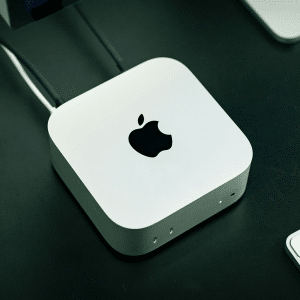 A silver Apple Mac Mini computer is placed on a black desk, connected by two cables at the back. Nearby, part of a white keyboard and another device can be seen—perfect for comparing internal vs external storage for your Mac Studio setup.