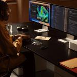 A person with long hair, glasses, and a yellow jacket sits at a desk using a keyboard, working on two large monitors displaying code and graphics. Office items and the new Mac Studio are nearby, perfectly set up for moving files in a seamless transition.