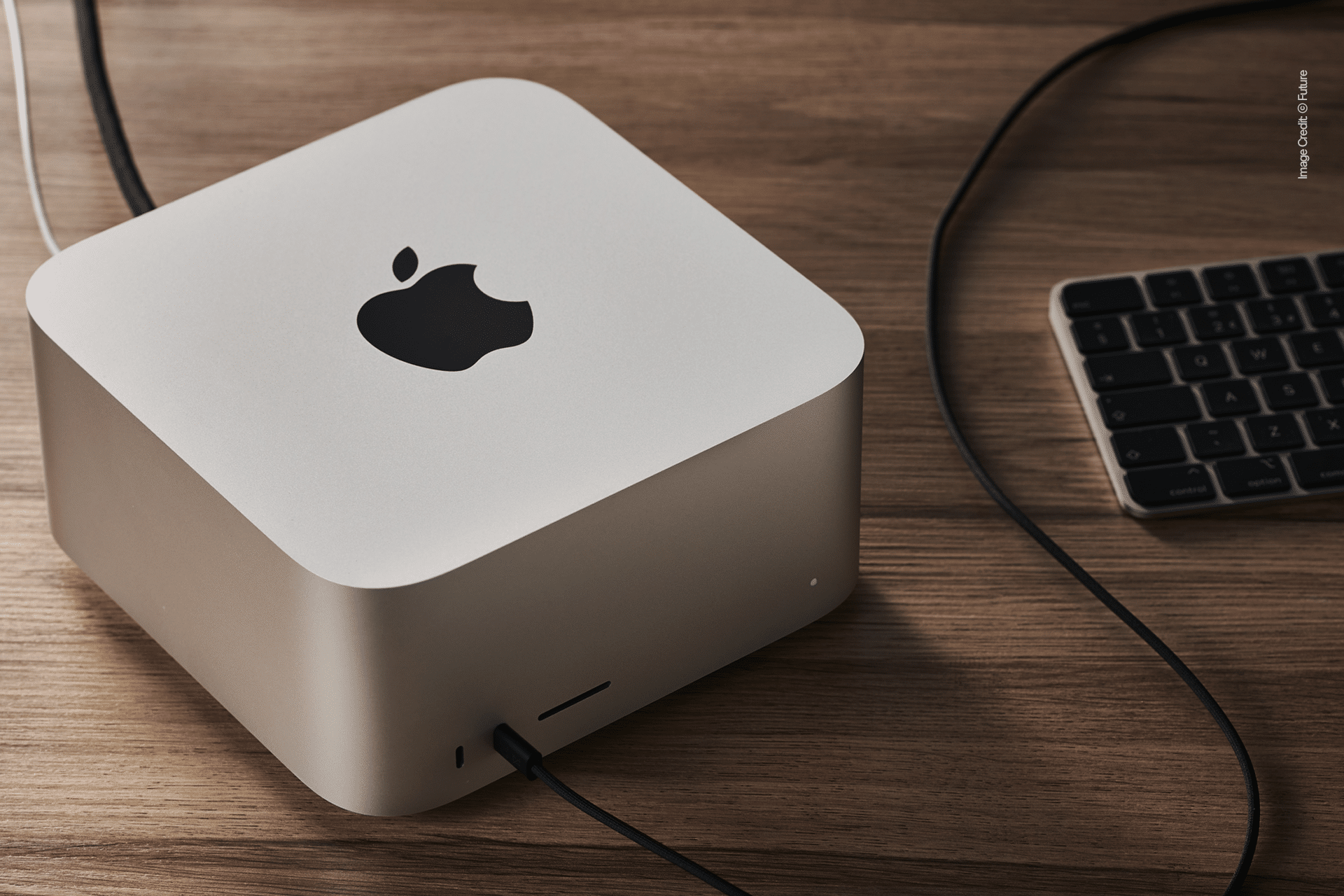 A silver Mac Studio desktop computer sits on a wooden desk next to a black keyboard, with cables connected to the ports—perfect for managing your workflow or automating tasks using the Shortcuts App.