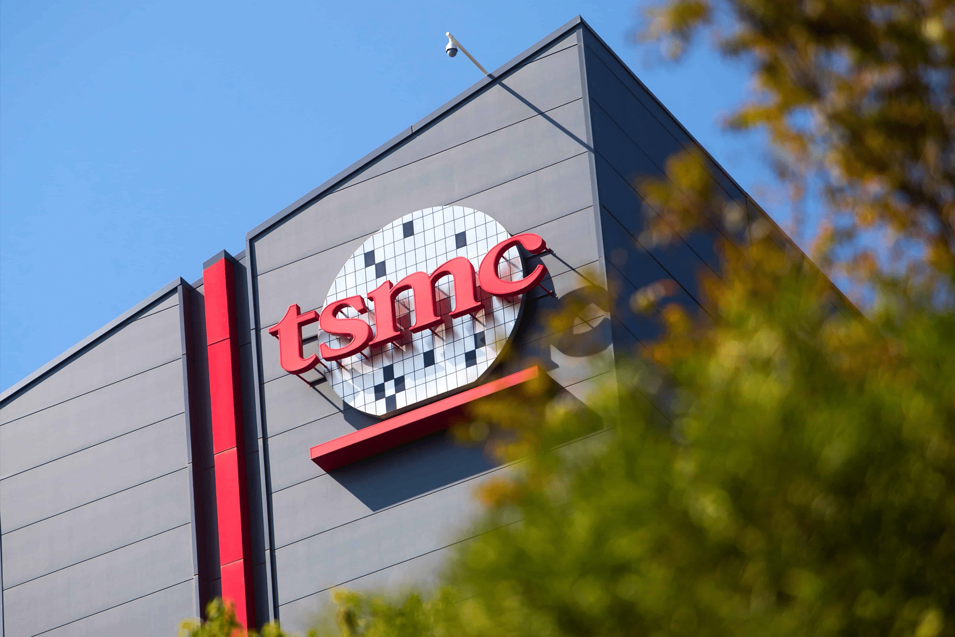 The TSMC logo is displayed on the side of a modern gray building, key to the global chips industry and partners like Nvidia, with a blue sky in the background and some green foliage in the foreground.