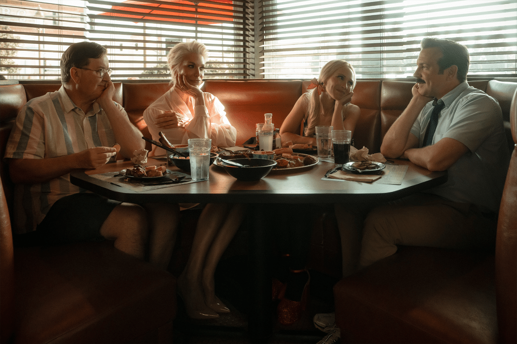 Four people sit in a dimly lit diner booth, facing each other across a table filled with food and drinks. Warm light filters through blinds behind them, evoking the cozy, nostalgic atmosphere of a classic Ted Lasso moment.