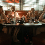 Four people sit in a dimly lit diner booth, facing each other across a table filled with food and drinks. Warm light filters through blinds behind them, evoking the cozy, nostalgic atmosphere of a classic Ted Lasso moment.