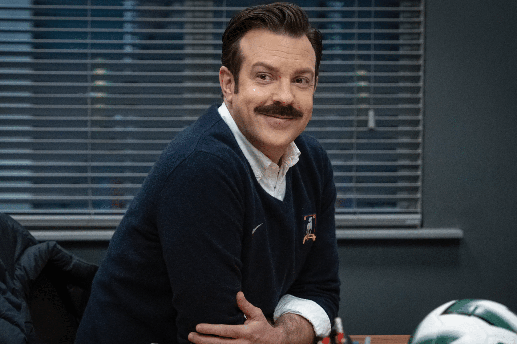 A man with brown hair and a mustache, resembling Ted Lasso, sits at a desk smiling in a navy sweater over a white shirt. Closed blinds are behind him and a soccer ball rests on the desk.