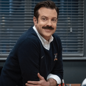A man with brown hair and a mustache, resembling Ted Lasso, sits at a desk smiling in a navy sweater over a white shirt. Closed blinds are behind him and a soccer ball rests on the desk.