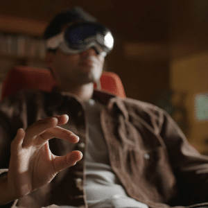 A person wearing VR goggles sits in a chair, making an "OK" gesture with their hand. The softly blurred background features shelves and warm lighting, creating a relaxed atmosphere that hints at visionOS accessibility and advanced eye support.