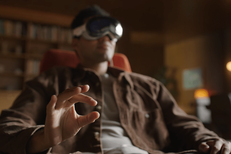 A person wearing VR goggles sits in a chair, making an "OK" gesture with their hand. The softly blurred background features shelves and warm lighting, creating a relaxed atmosphere that hints at visionOS accessibility and advanced eye support.