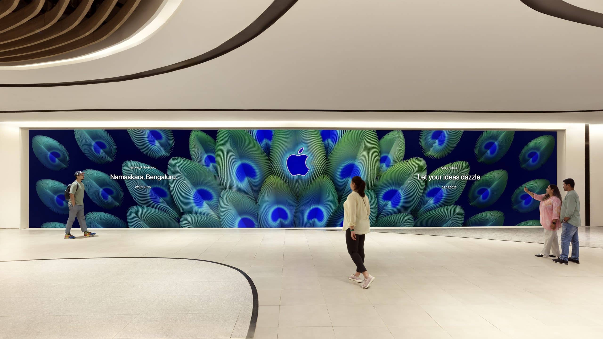 A large Apple Store mural with a peacock feather design and the Apple logo is displayed on a wall at the new Apple Hebbal Store in Bengaluru. Four people walk by, and text reads, “Namaskara, Bengaluru” and “Let your tech ideas dazzle.”.