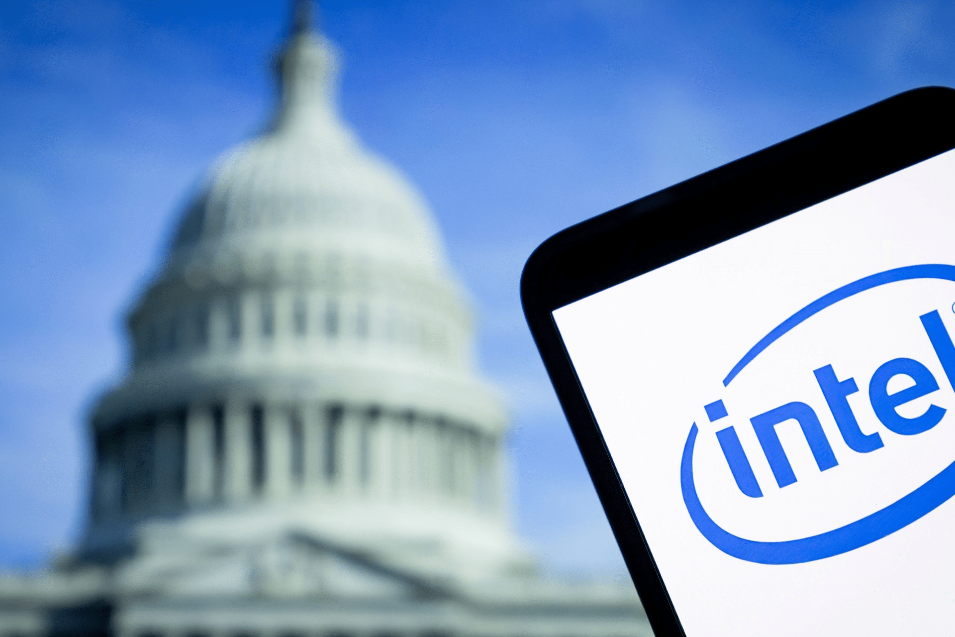 A smartphone displays the Intel logo in front of the blurred dome of the United States Capitol building, highlighting Intel's chip manufacturing ambitions in Ohio against a blue sky.