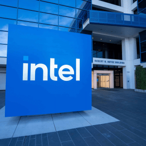 A large blue Intel logo stands in front of the glass entrance to the modern Robert N. Noyce Building, highlighting Intel’s leadership in chip manufacturing. The scene is outdoors, with some greenery visible on the right and a nod to Intel's Ohio presence.