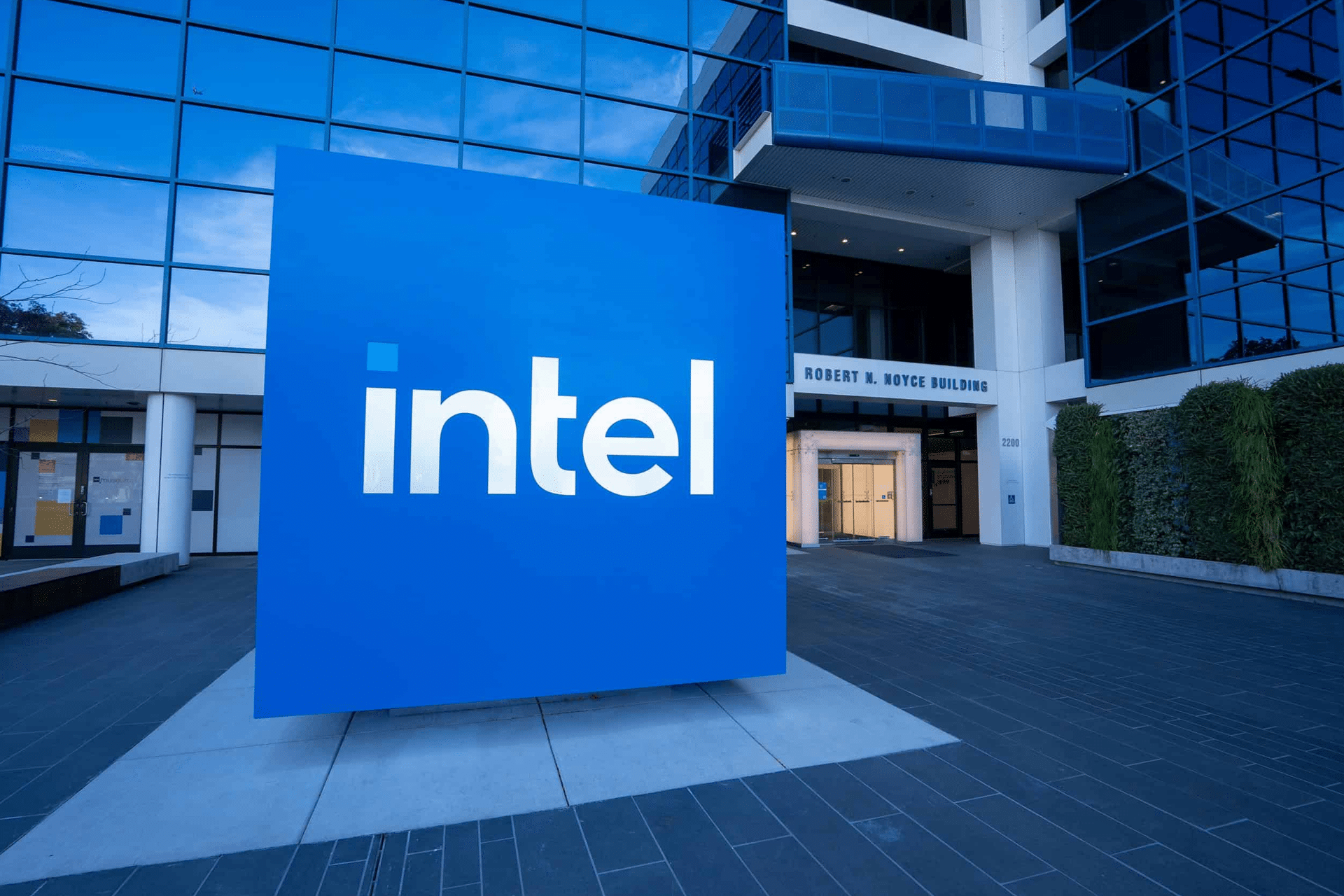 A large blue Intel logo stands in front of the glass entrance to the modern Robert N. Noyce Building, highlighting Intel’s leadership in chip manufacturing. The scene is outdoors, with some greenery visible on the right and a nod to Intel's Ohio presence.