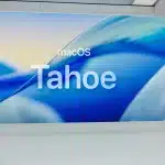 A person stands in front of a large screen displaying “macOS Tahoe Public Beta” with a blue abstract background in a modern, bright room, capturing the excitement of the latest innovation in Mac Computing.