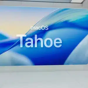 A person stands in front of a large screen displaying “macOS Tahoe Public Beta” with a blue abstract background in a modern, bright room, capturing the excitement of the latest innovation in Mac Computing.