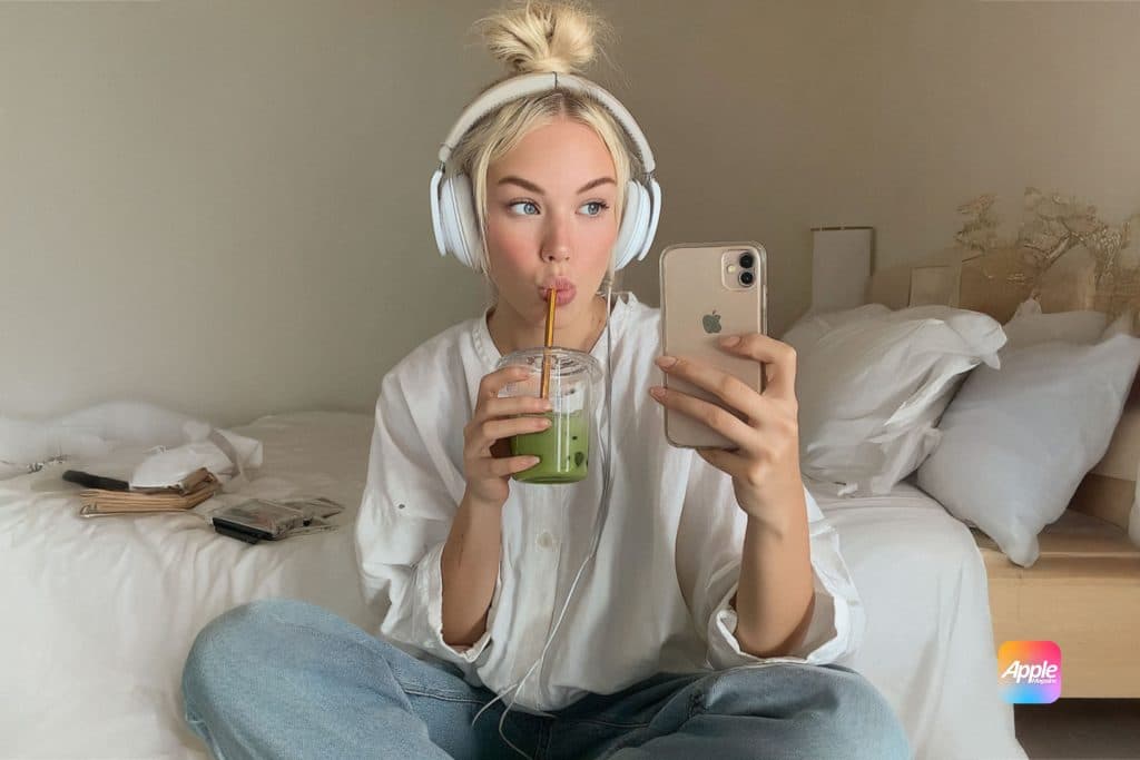A young woman with headphones sits cross-legged on a bed, sipping a green drink from a jar and taking a selfie with her phone. Perfect for content creators, she wears a white shirt and jeans. The Apple logo is visible in the corner.