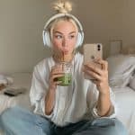 A young woman with headphones sits cross-legged on a bed, sipping a green drink from a jar and taking a selfie with her phone. Perfect for content creators, she wears a white shirt and jeans. The Apple logo is visible in the corner.