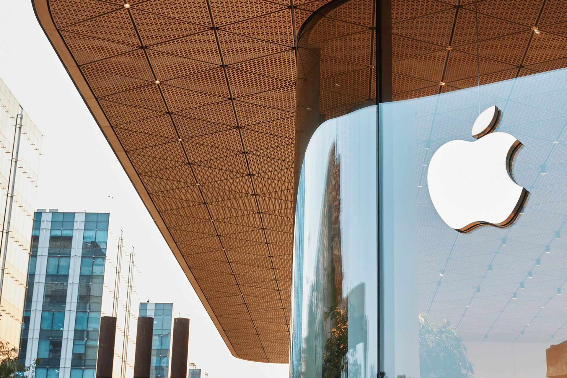 The photo shows the modern Apple Hebbal Store with a large glass facade and a prominent white Apple logo, set beneath a geometric-patterned roof, showcasing tech in Bengaluru amid tall buildings in the background.