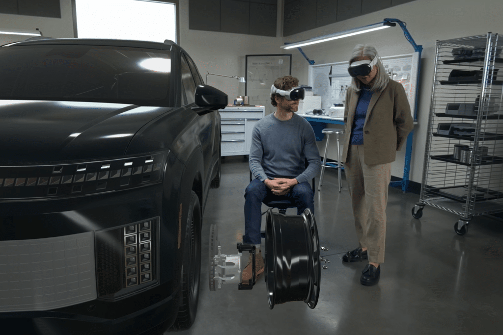 Two people wearing VR headsets interact with a virtual car model and parts in a high-tech workshop, exploring 3D modeling and design workflows—with one person seated and the other standing—using visionOS technology.