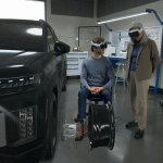 Two people wearing VR headsets interact with a virtual car model and parts in a high-tech workshop, exploring 3D modeling and design workflows—with one person seated and the other standing—using visionOS technology.