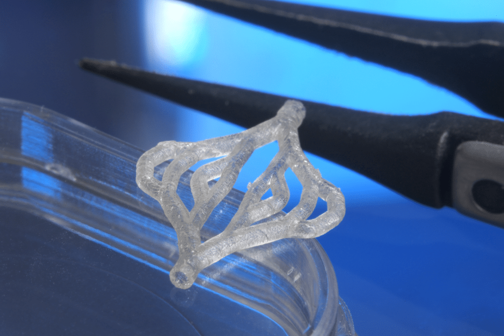 A close-up of a translucent, geometric 3D-printed artificial skin resting on the edge of a clear petri dish, with black tweezers in the background against a blue surface, highlighting innovation in regenerative medicine.