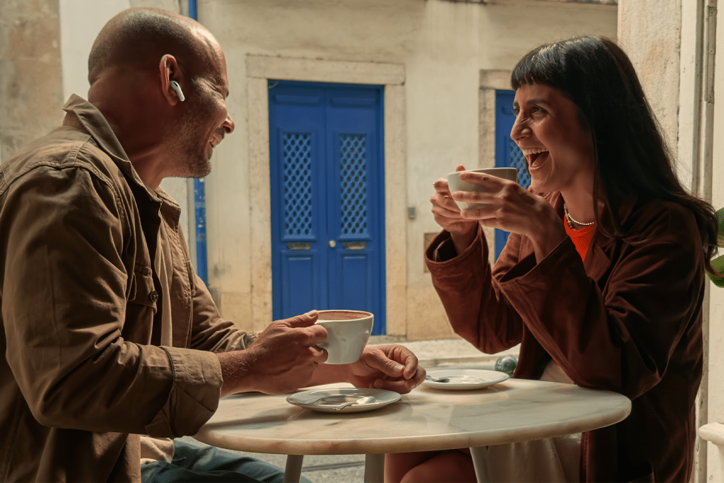 A man and a woman sit at an outdoor café table, laughing and holding coffee cups. On the table sits an AirPods Pro 3 case with a U2 chip, known for enhanced tracking. The background features white walls and bright blue doors and windows.