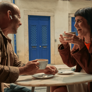 A man and a woman sit at an outdoor café table, laughing and holding coffee cups. On the table sits an AirPods Pro 3 case with a U2 chip, known for enhanced tracking. The background features white walls and bright blue doors and windows.