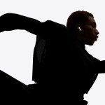A person with short red hair, wearing dark athletic clothing and Apple AirPods Pro 3 noise-canceling earbuds, is captured in a dramatic, dynamic pose that suggests running or dancing, arms and legs bent and silhouetted against a light background.