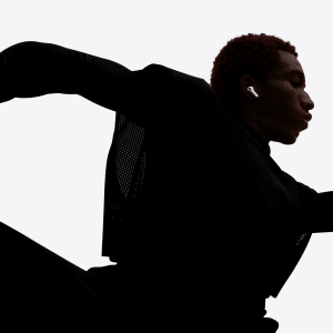 A person with short red hair, wearing dark athletic clothing and Apple AirPods Pro 3 noise-canceling earbuds, is captured in a dramatic, dynamic pose that suggests running or dancing, arms and legs bent and silhouetted against a light background.