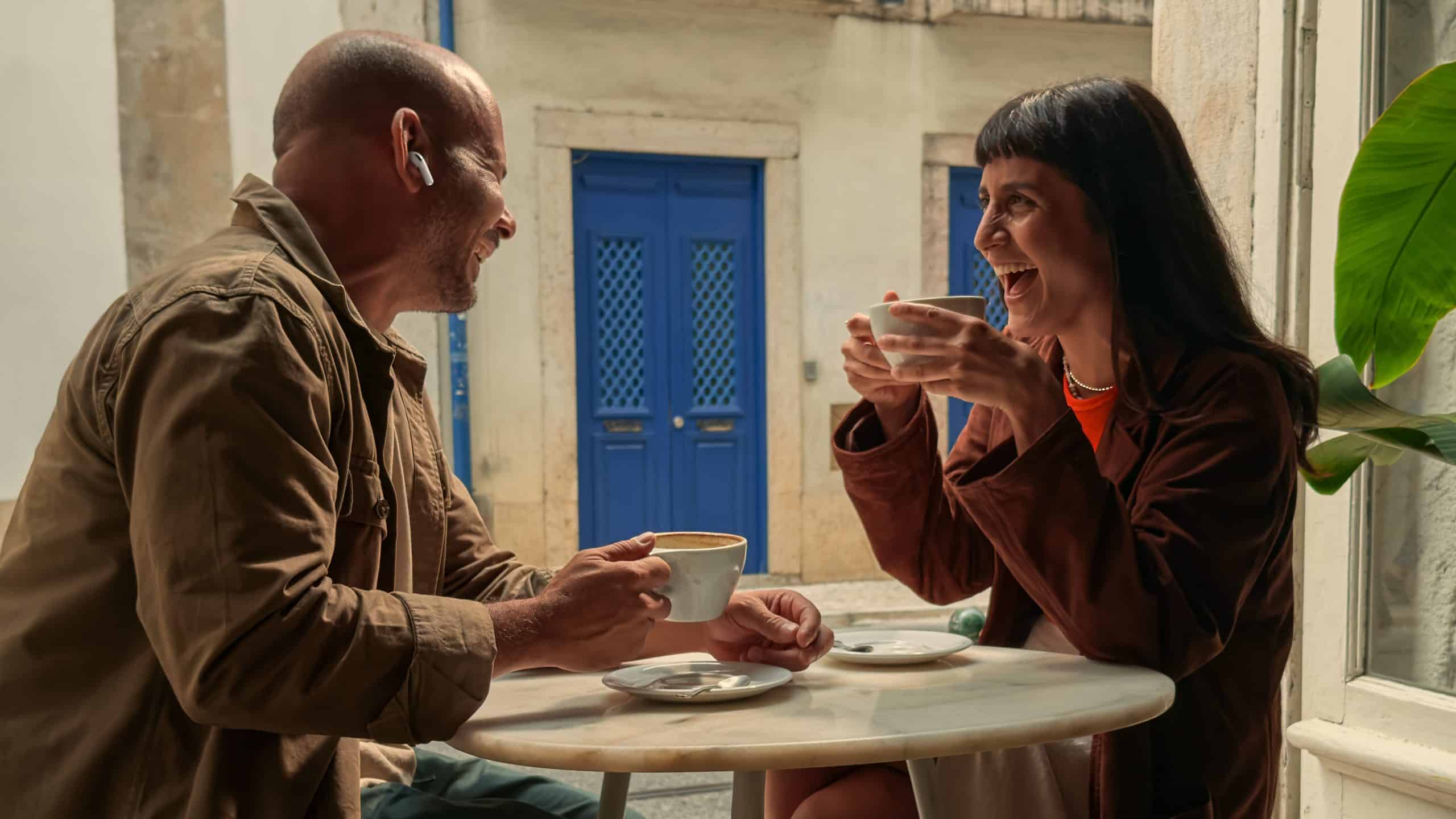 A man and woman sit at a small round table outside, smiling and laughing while holding coffee cups. As they chat using Live Language Translation with their AirPods Pro 3, blue doors and a sunlit building brighten the background.