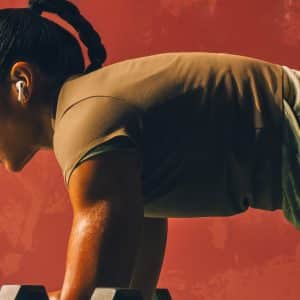 A woman with braided hair and AirPods Pro 3 holds a plank position on her forearms in a gym, sweat visible on her skin, against a reddish wall.