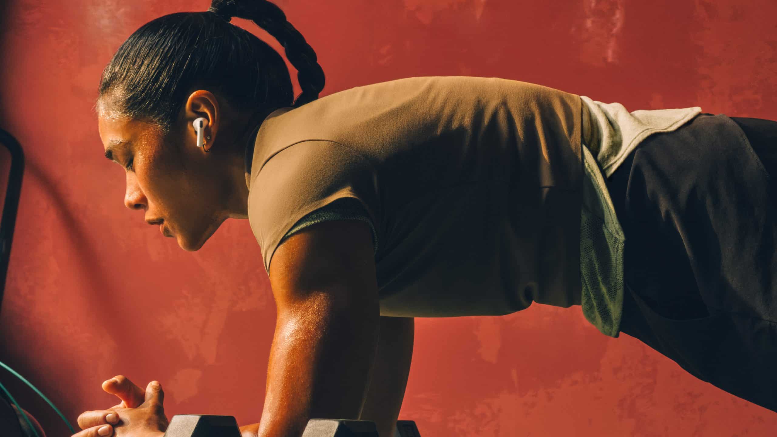 A woman with braided hair and AirPods Pro 3 holds a plank position on her forearms in a gym, sweat visible on her skin, against a reddish wall.