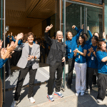 A group of people, many in matching blue shirts, stand and cheer outside the Apple Ginza store reopening, while three people in the front smile and wave to the crowd near the glass doors.