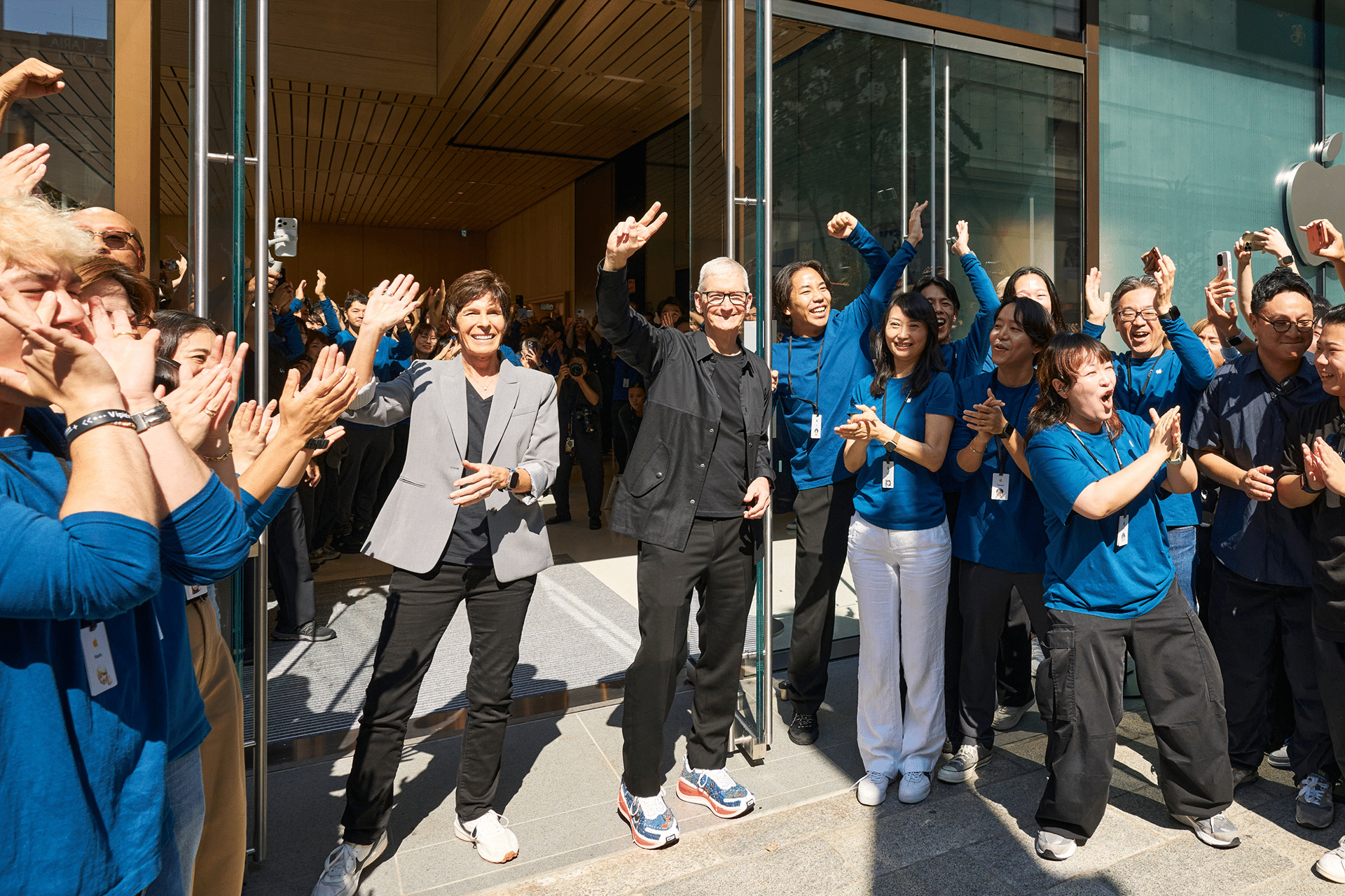 A group of people, many in matching blue shirts, stand and cheer outside the Apple Ginza store reopening, while three people in the front smile and wave to the crowd near the glass doors.