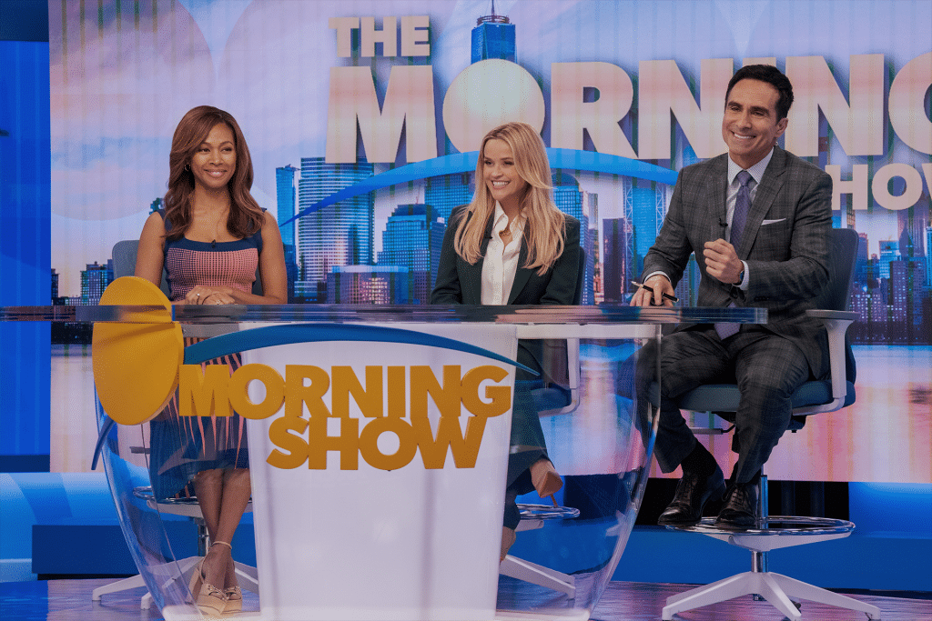 Three smiling TV hosts sit behind a glass desk on the brightly lit "The Morning Show" set, with a city skyline and large "Morning Show" text behind them—an exciting glimpse of Season 4 now streaming on Apple TV+.