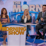 Three smiling TV hosts sit behind a glass desk on the brightly lit "The Morning Show" set, with a city skyline and large "Morning Show" text behind them—an exciting glimpse of Season 4 now streaming on Apple TV+.