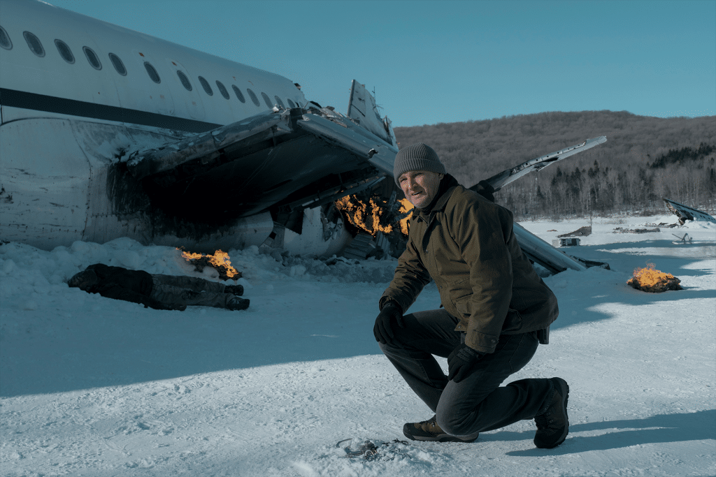 A man in winter clothing kneels in the snow near a crashed airplane with burning wreckage, surrounded by snow-covered trees and hills. Small fires mark the crash site as he faces a harsh survival challenge in Alaska's Last Frontier.