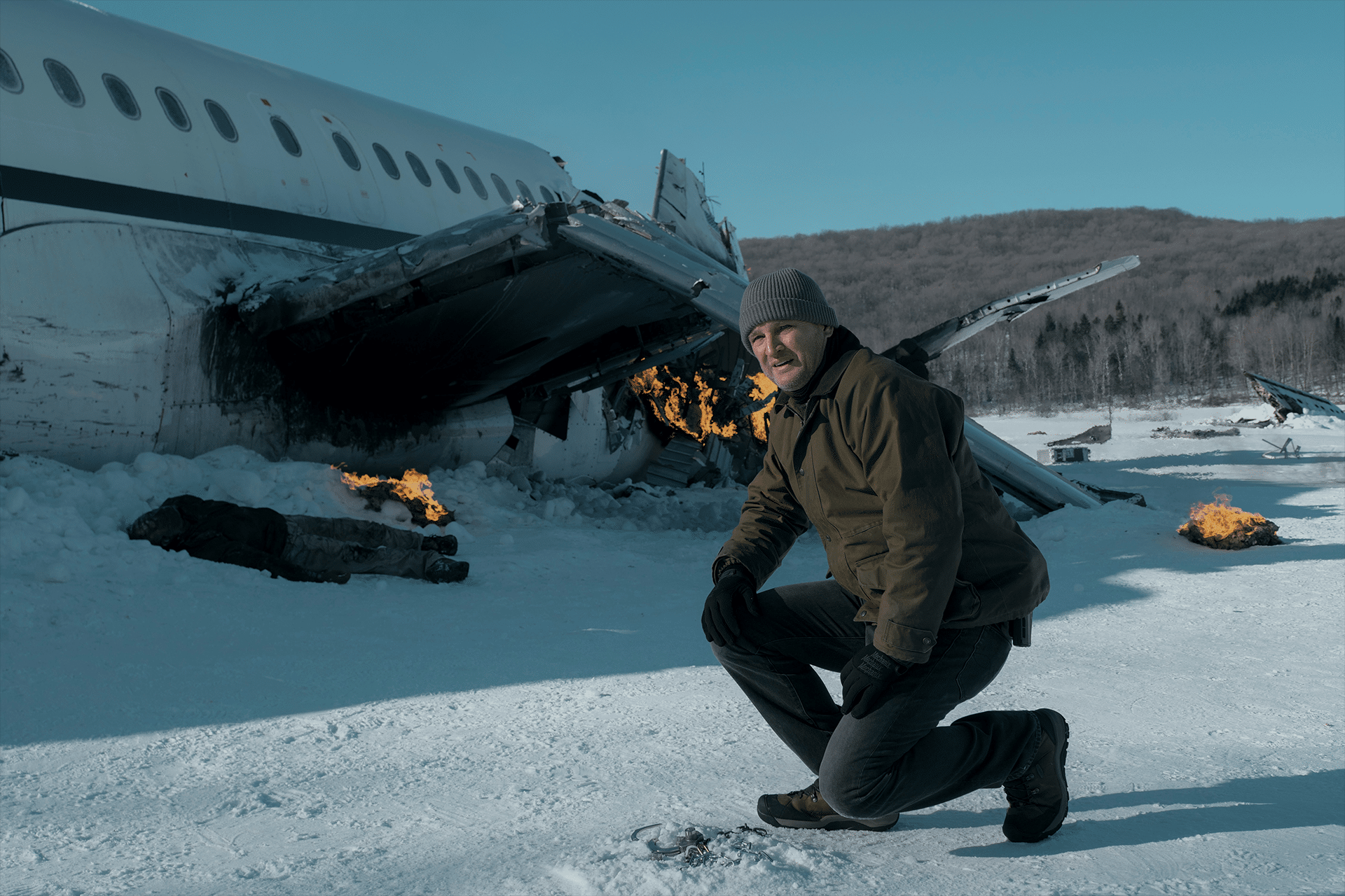 A man in winter clothing kneels in the snow near a crashed airplane with burning wreckage, surrounded by snow-covered trees and hills. Small fires mark the crash site as he faces a harsh survival challenge in Alaska's Last Frontier.