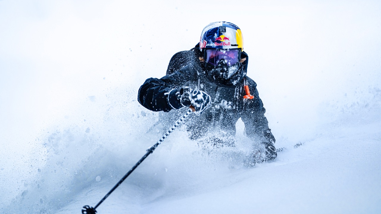 A skier in black gear and a helmet charges through deep powder snow, sending a spray into the air on a bright, wintry slope—perfect for experiencing immersive films with Vision Pro’s spatial storytelling. The skier's goggles reflect the snowy surroundings.