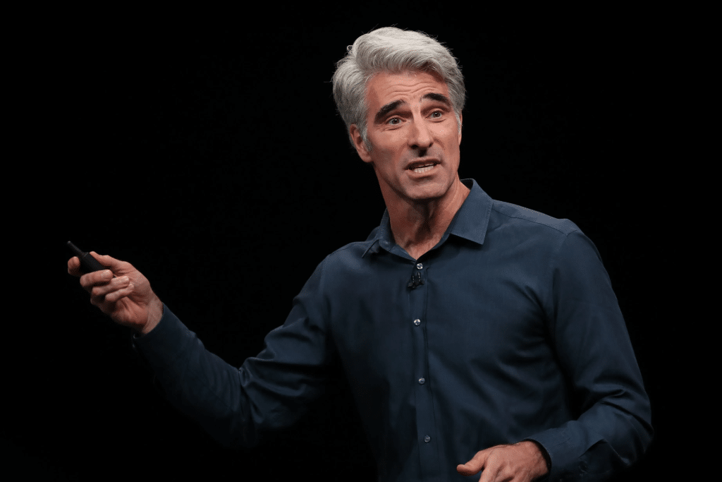 A man with gray hair wearing a dark blue shirt speaks animatedly on stage about the iPhone 17 Launch, holding a small device in his right hand, against a plain black background.