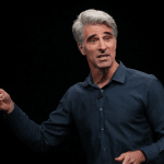 A man with gray hair wearing a dark blue shirt speaks animatedly on stage about the iPhone 17 Launch, holding a small device in his right hand, against a plain black background.