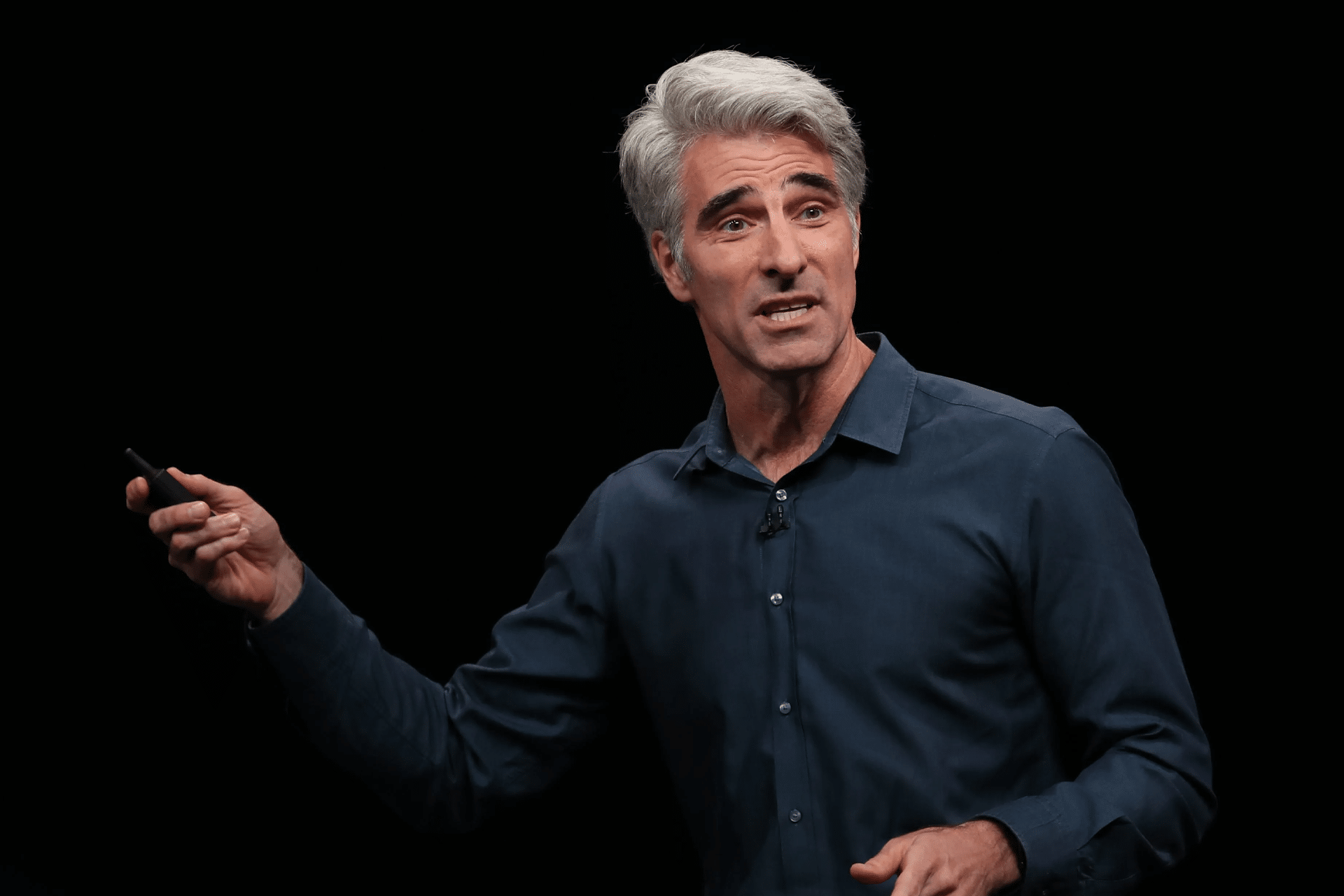 A man with gray hair wearing a dark blue shirt speaks animatedly on stage about the iPhone 17 Launch, holding a small device in his right hand, against a plain black background.
