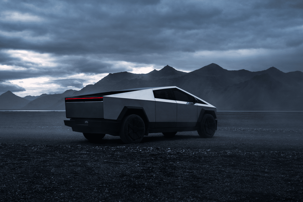 A futuristic, angular silver pickup truck equipped with Tesla Cybertruck modifications drives off-road on a dark, misty landscape with mountains in the background beneath a cloudy sky, ready for thrilling adventures.