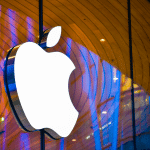 Large, illuminated Apple logo on the exterior of an Apple Store, set against a modern, curved wooden ceiling and reflective glass windows—a striking symbol amid discussions on the Digital Markets Act.