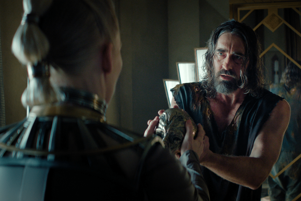 A disheveled man in dark, tattered clothing holds a detailed artifact while facing a white-blonde figure in ornate metallic attire inside a modern, dimly lit room—a striking scene from Foundation renewed on Apple TV+.