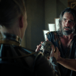 A disheveled man in dark, tattered clothing holds a detailed artifact while facing a white-blonde figure in ornate metallic attire inside a modern, dimly lit room—a striking scene from Foundation renewed on Apple TV+.