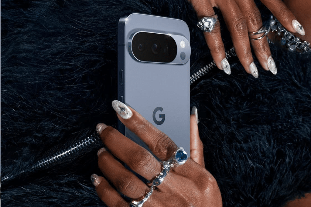 A person with manicured nails and multiple rings holds a blue Google Pixel smartphone against a black, furry textured background—perfect for Pixel users eager to explore the latest Android features.