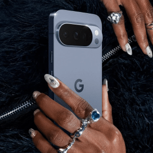 A person with manicured nails and multiple rings holds a blue Google Pixel smartphone against a black, furry textured background—perfect for Pixel users eager to explore the latest Android features.